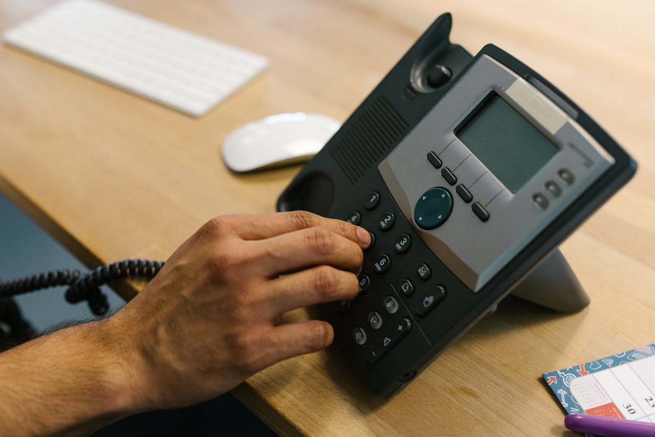 AI革新客服體驗:查詢,報Fault冇難度! A hand dialing a phone on a wooden desk. Office environment with keyboard and planner.